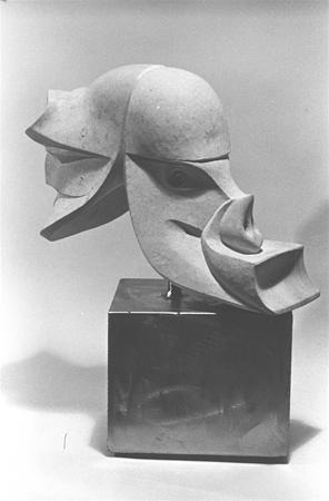 Abstract sculpture of a stylized head with geometric shapes and curves, mounted on a rectangular base. The artwork features overlapping forms and smooth surfaces, creating a modern and artistic appearance. Black and white photo.