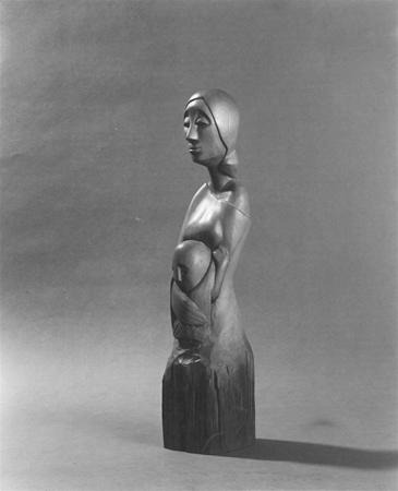 A wooden sculpture of a stylized, elongated female figure with a serene expression, arms folded, and a rounded belly, standing on a rough, uncarved base against a plain background.
