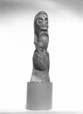 A wooden sculpture depicting a bearded figure with elongated features, holding a smaller figure close to its chest, displayed on a cylindrical pedestal against a plain background.