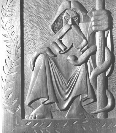 A carved wooden relief depicts a stern, hooded figure sitting, gripping a staff entwined with a snake, with stylized leaves bordering the left side. The figure’s expression is intense and commanding.