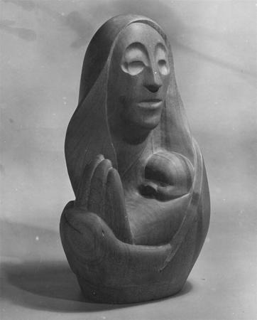 A smooth, carved wooden sculpture depicts a stylized figure of a woman in a hooded cloak cradling a baby in her arms. The figures have simple, abstract facial features. The background is plain and softly lit.