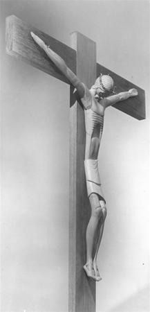 A black-and-white photo of a wooden crucifix featuring a stylized, elongated figure of Jesus Christ with arms outstretched, wearing a cloth around the waist. The background is plain and light-colored.