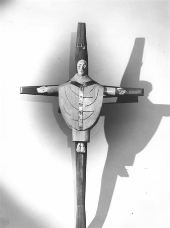 A wooden crucifix featuring a stylized, rounded figure with outstretched arms, a calm facial expression, carved garment details, and highlighted hands and feet, casting a shadow on a plain background.