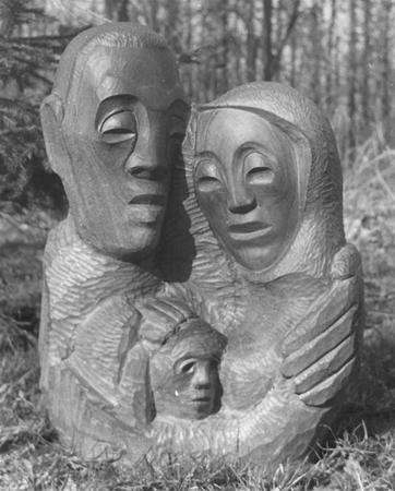 A wooden sculpture depicting three abstract human figures, with two larger figures embracing a smaller figure, set outdoors with trees in the background.