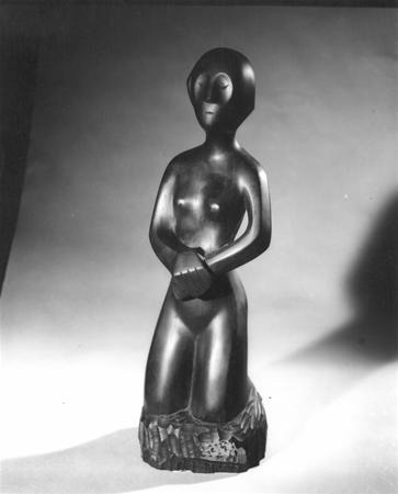 A smooth, stylized sculpture of a nude, kneeling human figure with minimal facial features and arms crossed, set against a plain background. The lower part of the figure blends into a textured base.