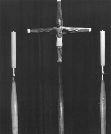 Black and white photo of a crucifix with a figure of Jesus in the center, flanked by two tall unlit candles on either side, against a dark background.