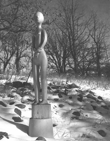 A smooth, abstract statue of a standing human figure is displayed on a pedestal outdoors in a snowy landscape, surrounded by leafless trees and scattered rocks.