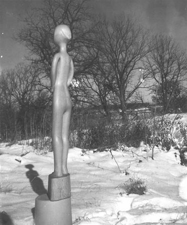 A smooth, abstract statue of a standing human figure on a pedestal is set outdoors in a snowy field, with leafless trees and a cloudy sky in the background.