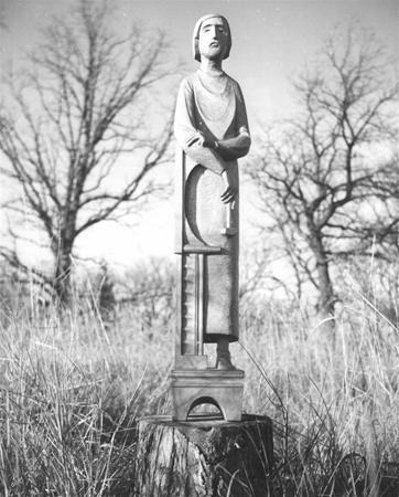 Black and white photo of a stylized, elongated statue of a person standing on a tree stump in a grassy field with leafless trees in the background. The statue has an abstract, geometric design.