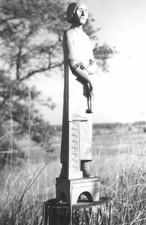 Black and white photo of a tall, slender sculpture of a person in a robe, standing on a pedestal outdoors in a grassy field, with blurred trees and sky in the background.