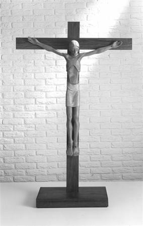 A wooden crucifix with a stylized figure of Jesus Christ is displayed against a white brick wall, with light casting soft shadows on the background.