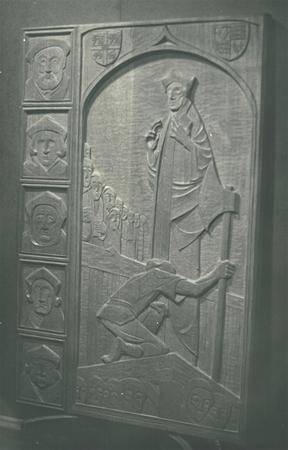 A carved wooden panel shows a large robed figure holding a staff, with a kneeling person in the foreground. To the left, smaller sculpted faces are arranged vertically in separate sections. The scene has a medieval style.