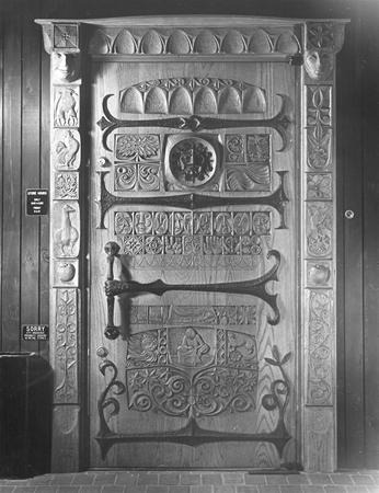 A large, ornate wooden door with intricate carvings of patterns, faces, and symbols, featuring a heavy metal handle and decorative panels, set in a dark wood frame.