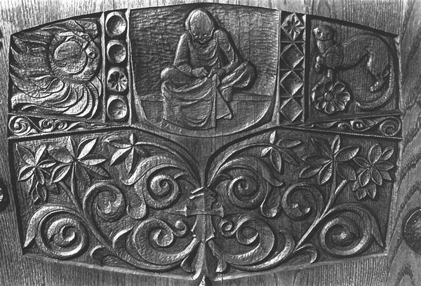 Detailed wood carving featuring a seated figure in the center, surrounded by floral patterns, a bird on the left, and an animal resembling a lion or dog on the right. Patterns and textures fill the background.