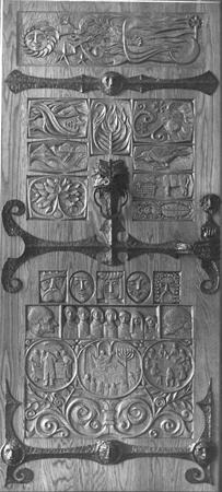 Ornate wooden door with intricate carvings, including floral patterns, human figures, and decorative metalwork. The reliefs are arranged in panels, showcasing scenes and symbols in a detailed, artistic style.