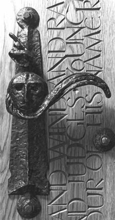 A metal door handle shaped like a face with an extended nose appears on a wooden door engraved with vertical and horizontal uppercase text.