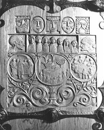 Carved wooden panel featuring portraits and scenes, including faces in medallions at the top and groups of people gathered around table-like structures with ornate swirling patterns below.