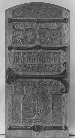 Black and white photo of a wooden door with ornate carvings, metal hinges, and the words "BRYDE WOOD" at the top. Decorative patterns and two faces are carved above nature-themed panels.