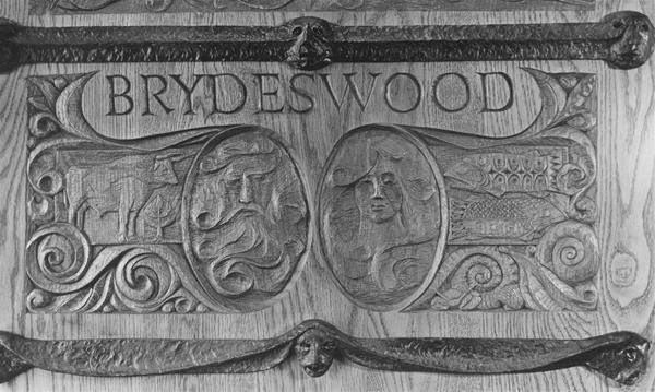 Carved wooden panel with the word "BRYDESWOOD" above two oval designs, one showing a lion and a castle, the other a woman's face and fish, surrounded by ornate patterns and animal faces on horizontal bars.