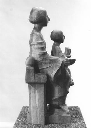 Wooden sculpture of a seated woman holding a book, with a child sitting on her lap. Both figures have stylized, simplified features. The sculpture rests on a patterned base.