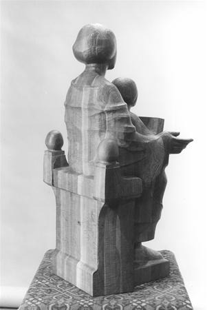 A wooden sculpture of a seated figure holding a child, viewed from behind. The figures sit on a throne-like chair with decorative spheres on the armrests, and the base features a patterned design.