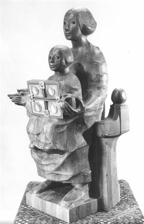 Wooden sculpture of two seated figures, one adult and one child, with the child holding a square object decorated with circles. Both figures have simple, stylized features and wear draped garments.