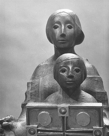 A wooden sculpture of two stylized figures, one adult and one child, both with simplified, rounded facial features and large eyes, positioned closely together with the child holding a rectangular object.