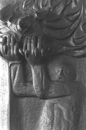 Carved wooden sculpture depicting a figure with upraised arms, holding or cradling a large, flowing organic form above their head. The figure's face is partially visible, appearing solemn or contemplative.