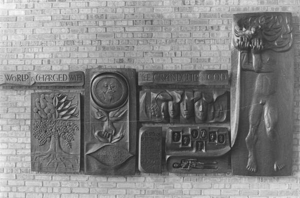 A relief sculpture on a brick wall depicts symbolic scenes, including a tree, a radiant star, several faces, and a tall standing figure. The words "The world is charged with the grandeur of God" are inscribed.