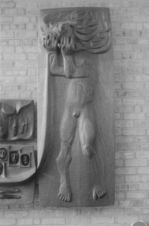 A wooden relief sculpture of a nude human figure standing against a brick wall, with hands raised to the face and flowing hair above the head, conveying an emotional or dramatic pose.