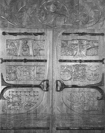 Black and white image of intricately carved wooden doors featuring segmented panels with various detailed figures, patterns, and scenes. A human figure with a raised hand is carved at the top center above the doors.