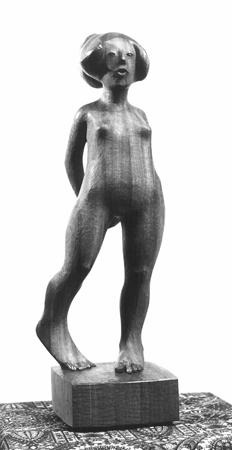A wooden sculpture of a nude, androgynous figure standing upright with hands behind the back. The figure stands on a rectangular base, with one knee slightly bent. The background is plain white.