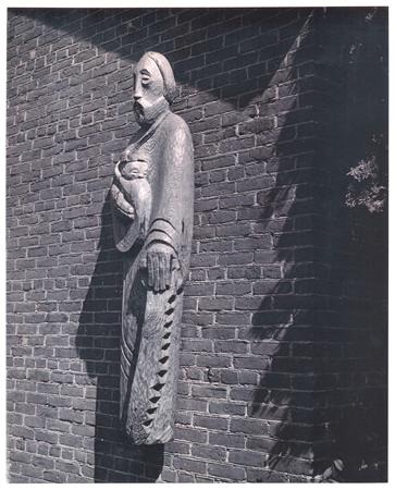 Wooden statue of a robed figure holding a child is mounted on a brick wall. The statue casts a shadow, and sunlight highlights the texture of the wall and the carved details of the figure.