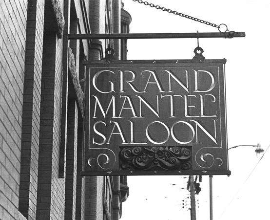 A hanging sign on a brick building reads “Grand Mantel Saloon” in bold, decorative letters, with a scrollwork design below the text.