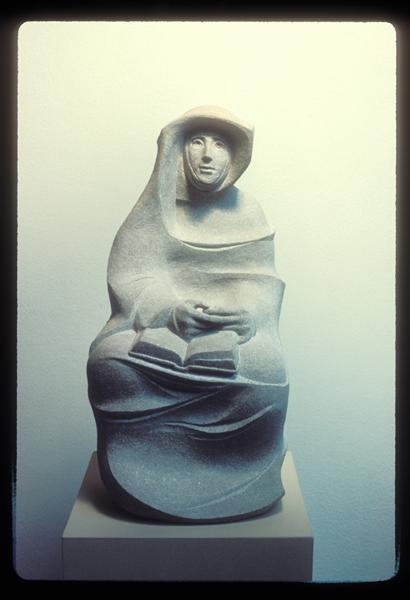 A stone sculpture of a seated, veiled figure holding an open book, displayed on a pedestal against a plain light-colored wall. The figure's features are stylized and abstract.