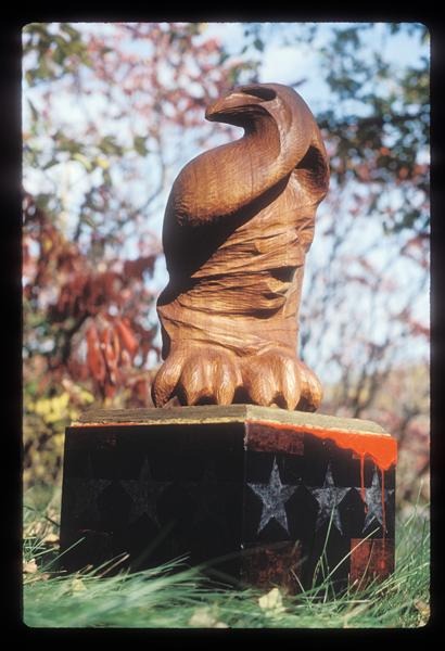A wooden eagle sculpture with stylized features sits atop a base decorated with white stars against a dark background. The scene is outdoors with blurred trees and foliage in the background.
