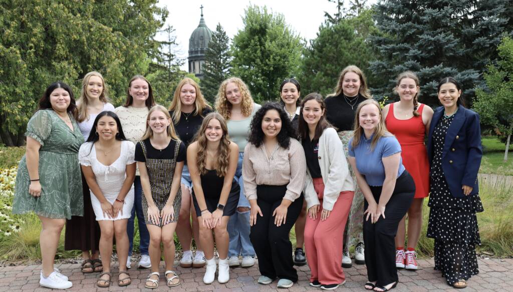 A group of fifteen young women pose for a photo outdoors in front of a lush garden with a historical building and trees in the background. They are all smiling and dressed in casual, summery attire, standing and crouching in two rows on a cobblestone path.