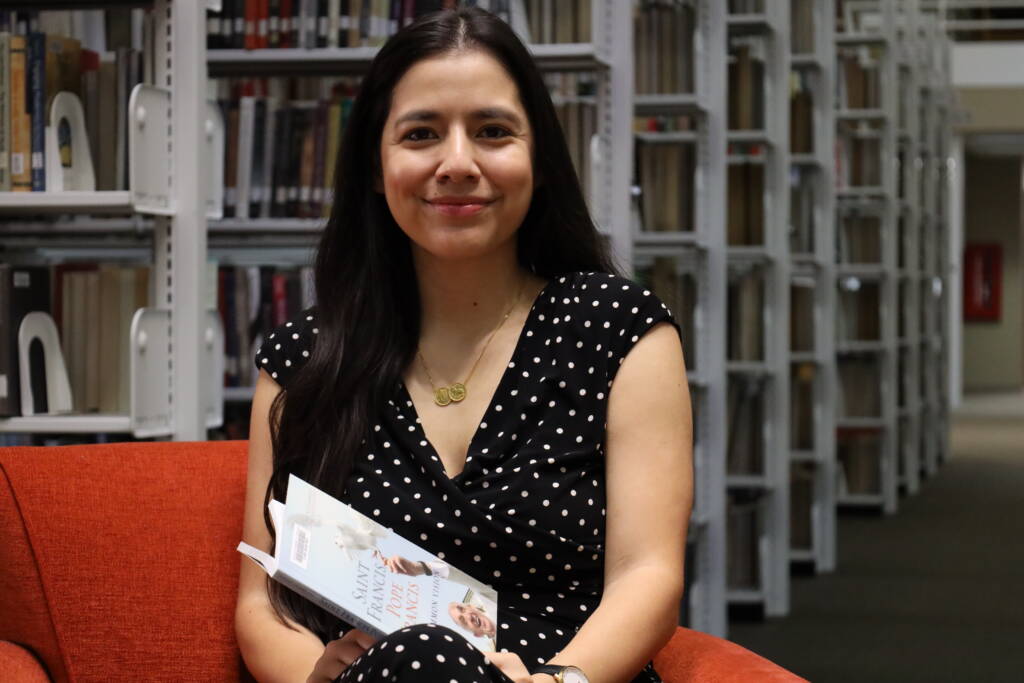 A woman with long dark hair, wearing a black dress with white polka dots, sits on an orange chair in a library, smiling and holding an open book. Shelves filled with books are visible in the background.