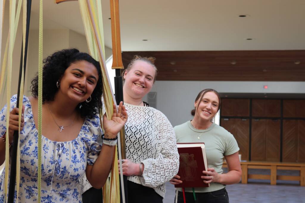 Three smiling women stand indoors; one holds ribbons, another wears a crocheted top, and the third holds a large red book. They appear relaxed and happy in a bright, open space with wooden accents.