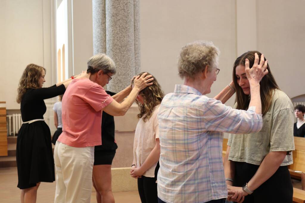 Three women stand in a church with heads bowed as three older women gently place their hands on their heads, appearing to bless or pray for them in a solemn ceremony.