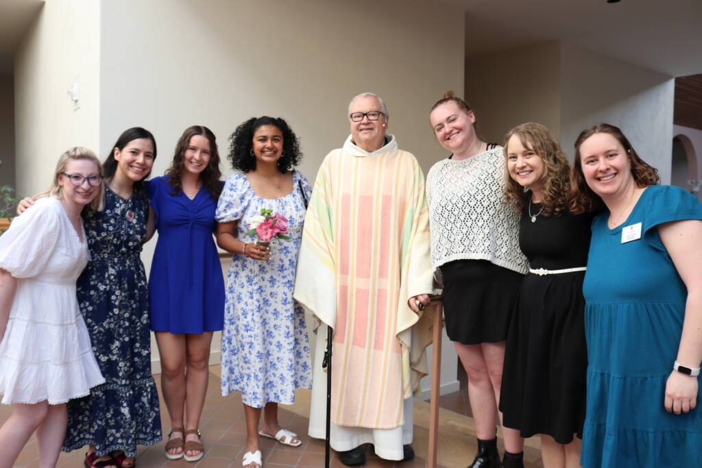 A group of seven smiling women and a priest in light-colored robes stand together indoors, posing for a photo. One woman in the center holds a bouquet of flowers. Everyone looks happy and dressed up.