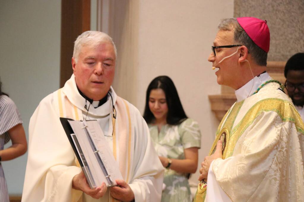 A priest holding a binder stands beside a bishop in ceremonial robes and a pink skullcap. They are indoors, with two people in the background, including a woman in a light green dress.