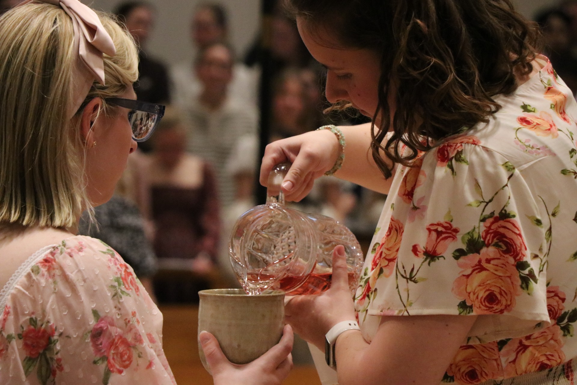 Two women in floral dresses participate in a ceremony; one pours a pink liquid from a glass pitcher into a cup held by the other. A group of people watch in the blurred background.