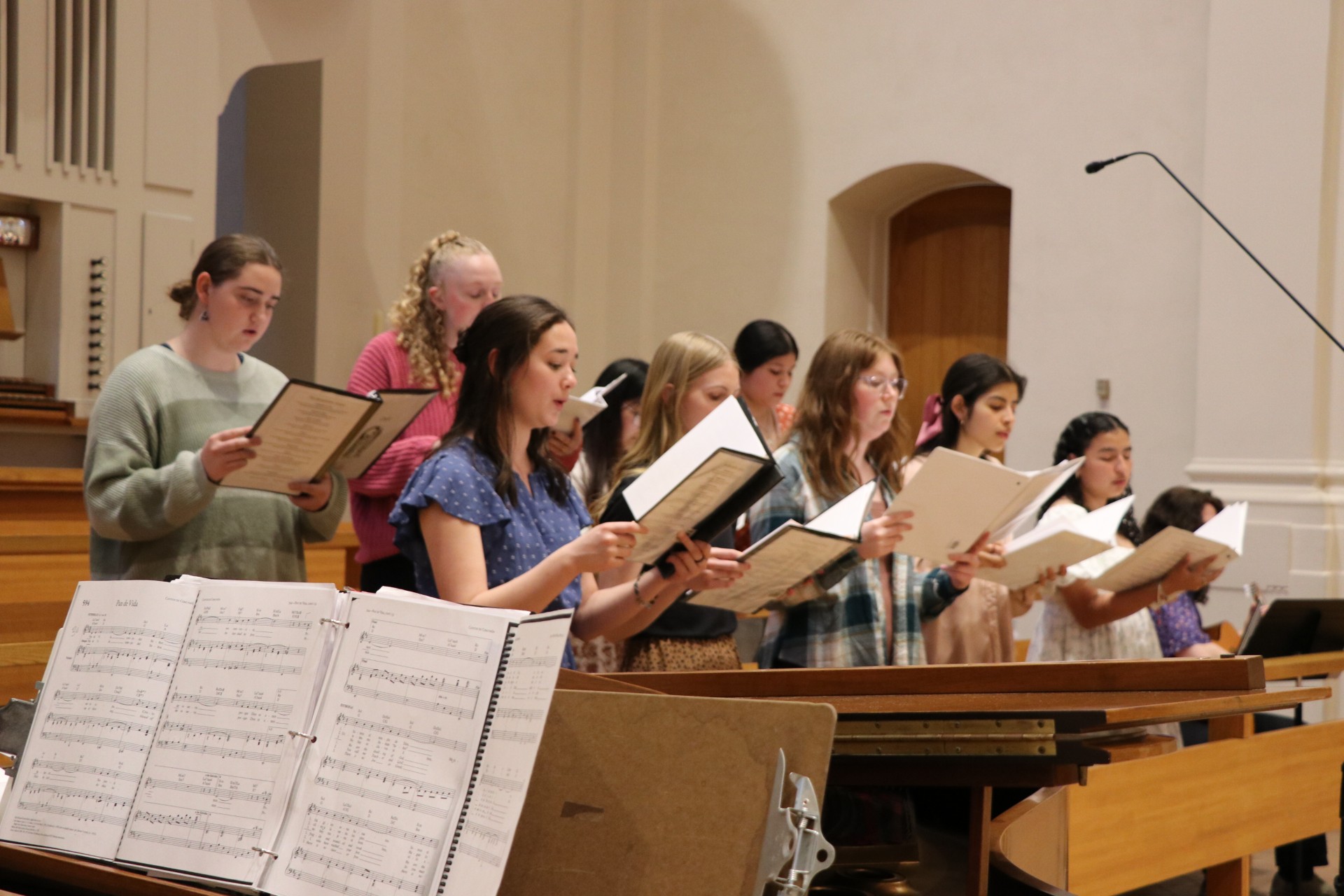 A group of young women standing in a church choir loft sing from sheet music, with songbooks open. Sheet music and musical stands are visible in the foreground. The setting is bright and spacious.