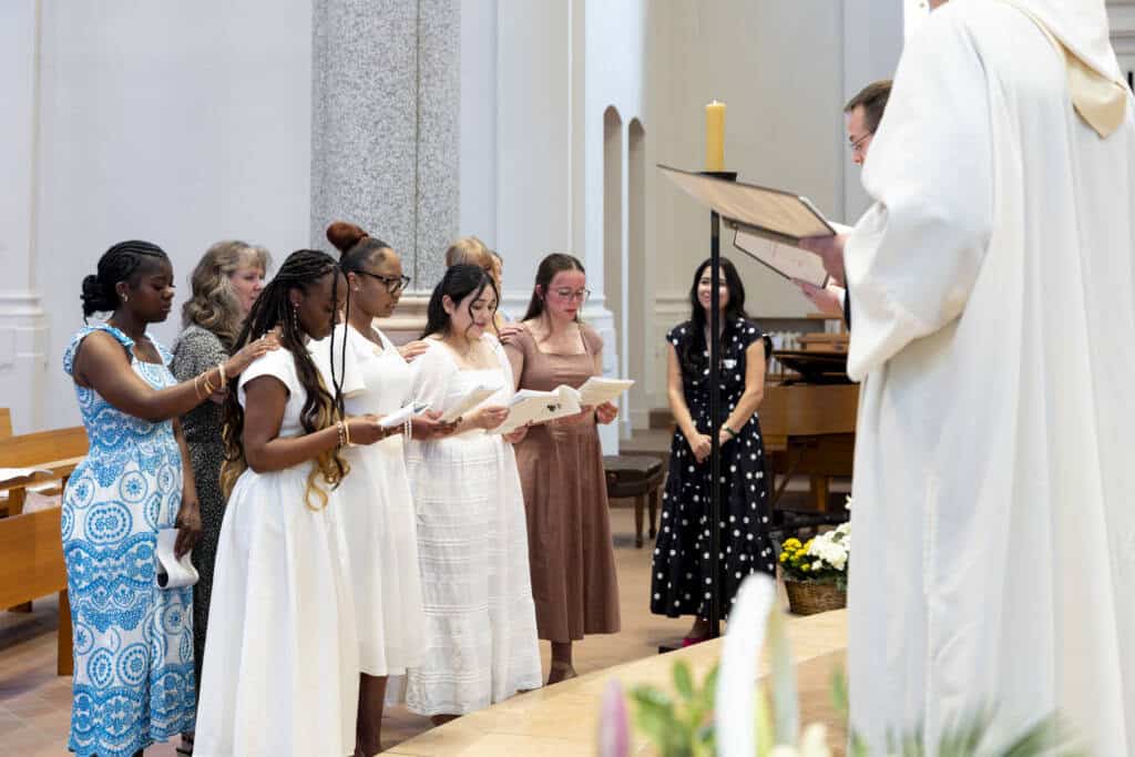 A group of young women in dresses stand in a church, holding papers and singing or reading, while a priest in white robes addresses them. One woman has her hand on another's shoulder in support.