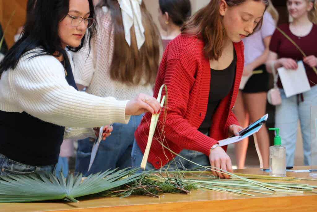 Two women arrange and weave palm leaves and greenery on a table. One holds a piece of paper and a phone, while others stand in the background. A bottle of hand sanitizer is also visible on the table.