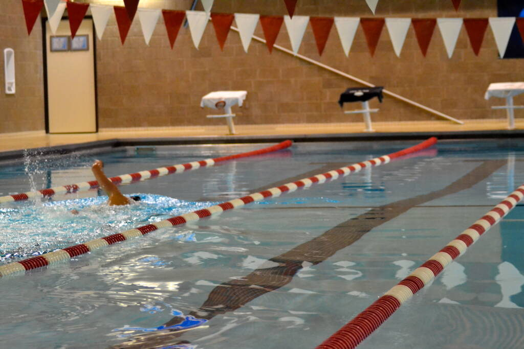 A swimmer performs a backstroke in an indoor pool, creating splashes in their lane. The pool is empty except for the swimmer; red-and-white lane dividers and triangular flags hang above the water.