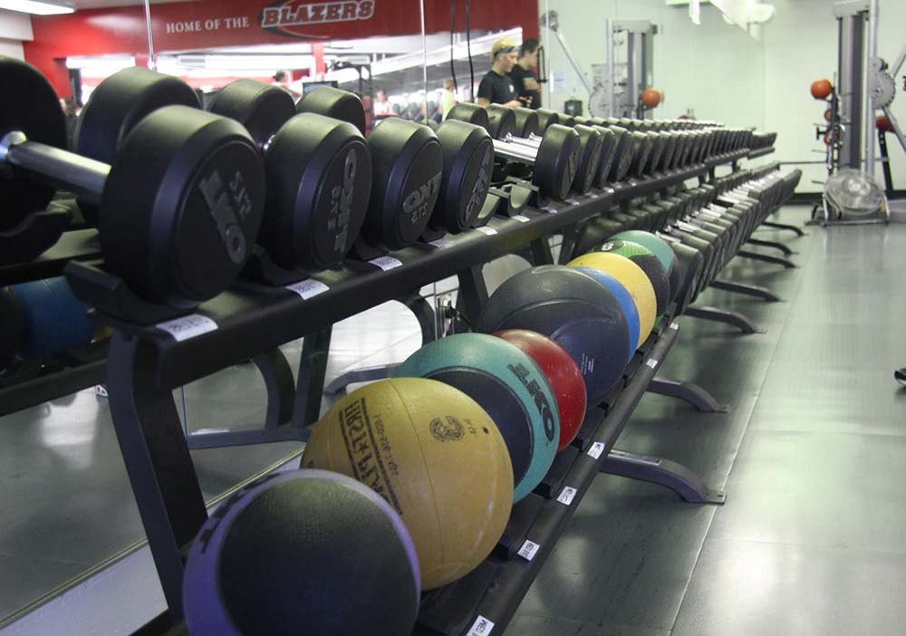 A row of dumbbells and colorful medicine balls are neatly arranged on racks in a gym, with strength training equipment and mirrors visible in the background.