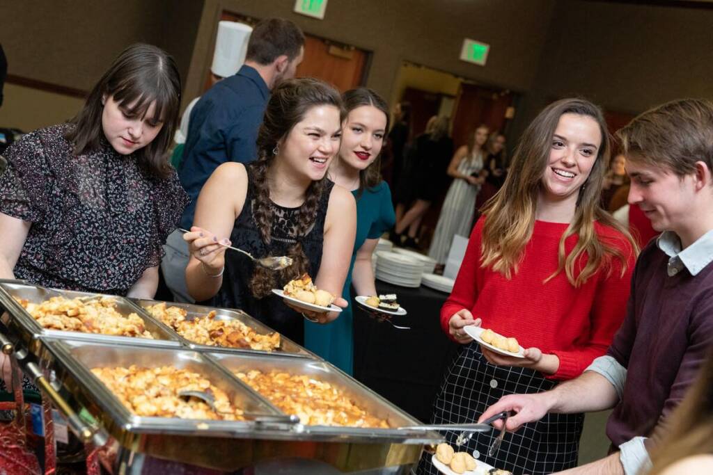 A group of young adults is gathered around a buffet table at a social event. They are serving themselves food from chafing dishes filled with pasta and other dishes. They are smiling and engaging in conversation, with a festive setting in the background.