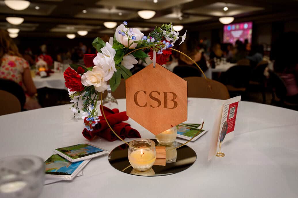 A table centerpiece with flowers, candles, and a wooden sign engraved with "CSB" sits on a round mirror at a banquet event. People are seated in the background under soft lighting.